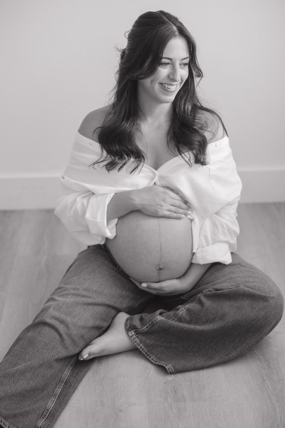 Black and white photo of a smiling pregnant woman sitting cross-legged on floor, cradling her baby bump in jeans and white top.