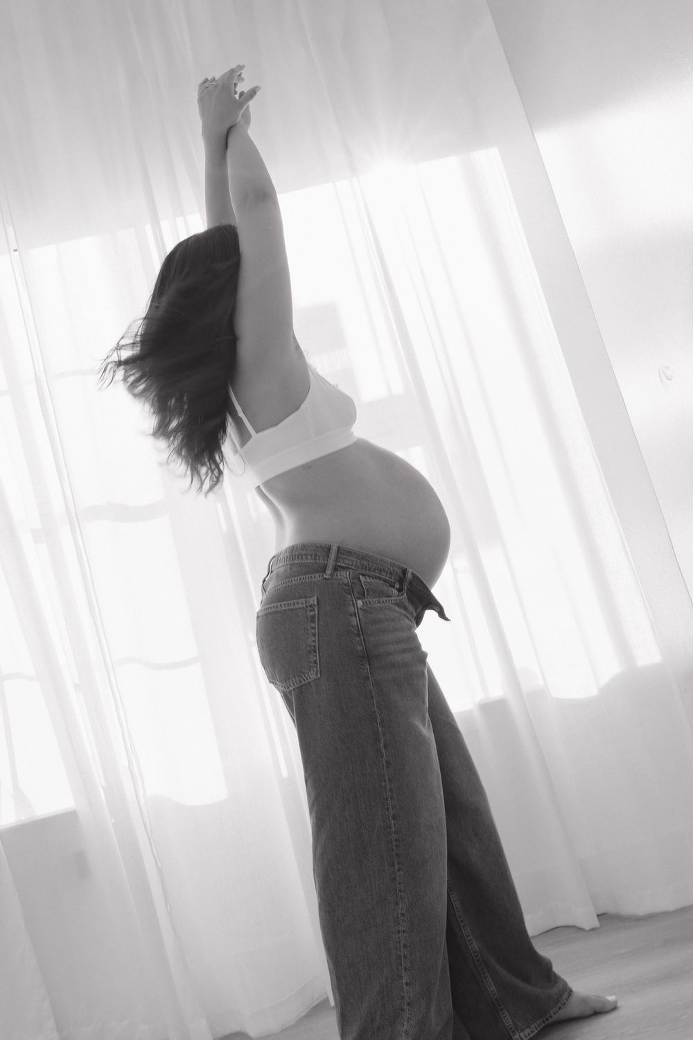 Black and white photo of a pregnant woman in jeans stretching her arms up near sheer white curtains.