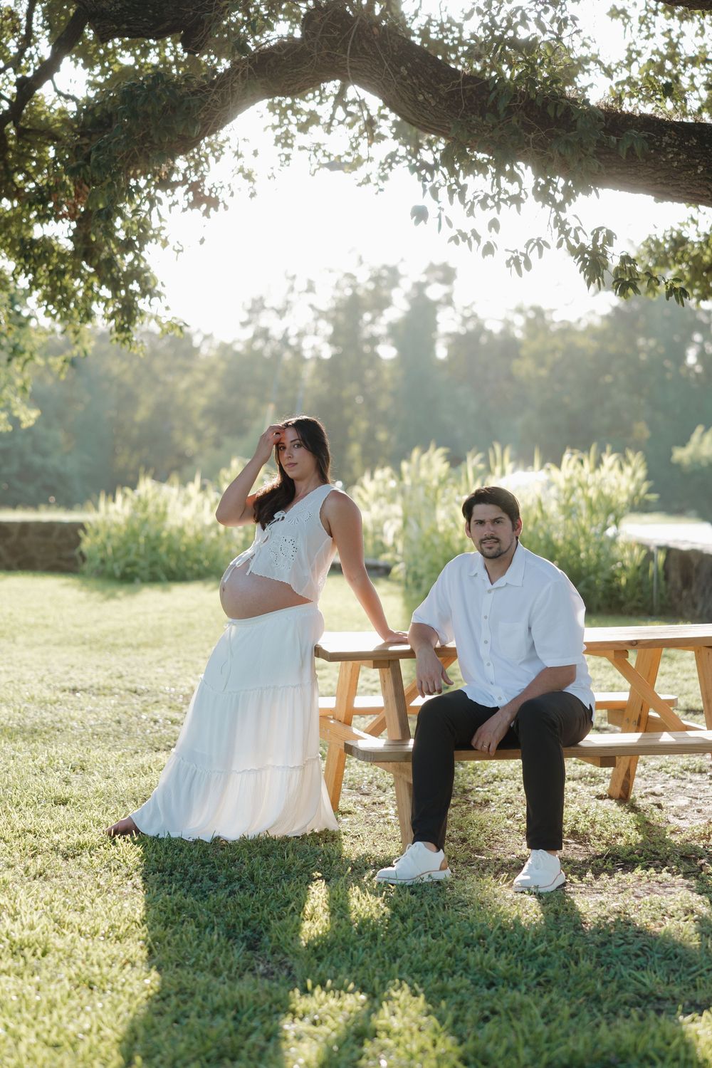 Pregnant woman in white dress poses with man at picnic table under tree in sunny park setting.