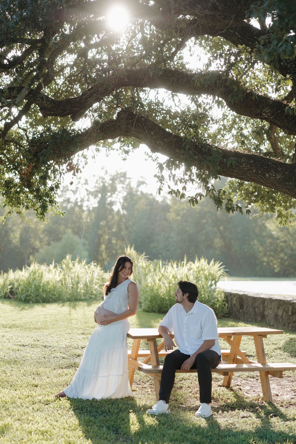 Pregnant woman in white dress stands under oak tree with partner at picnic table in sunny park setting.