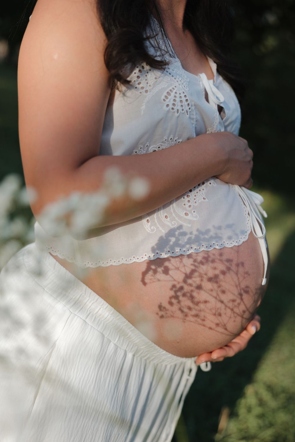 Pregnant woman in white lace top cradling bare baby bump outdoors with floral shadows in natural sunlight.