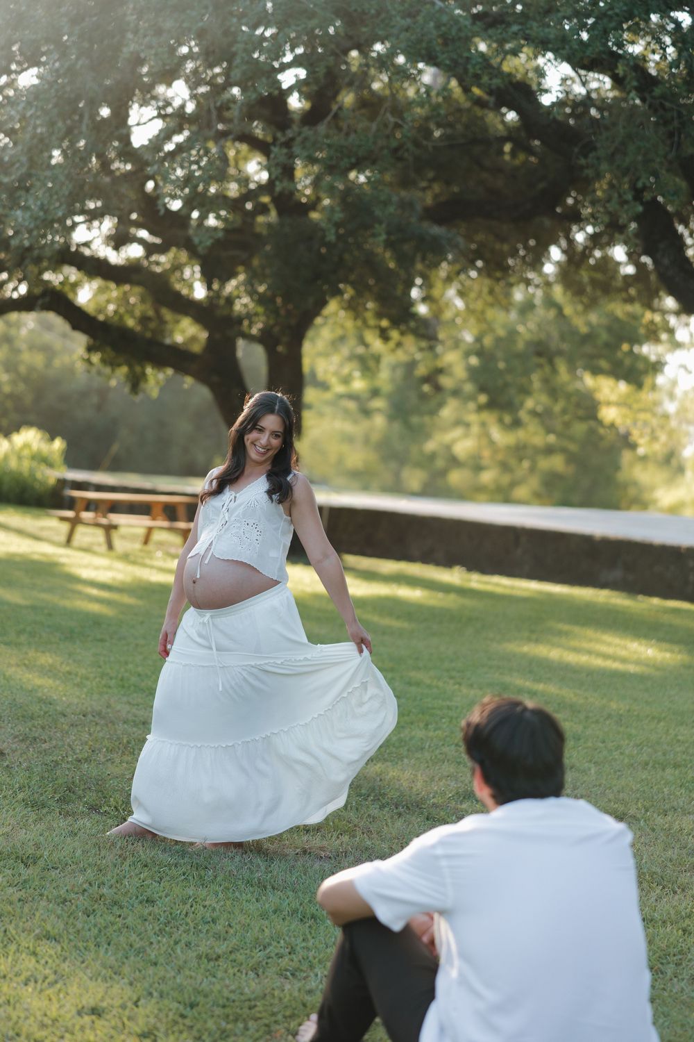 Pregnant woman in white dress holds her belly outdoors while partner watches during golden hour maternity photoshoot.