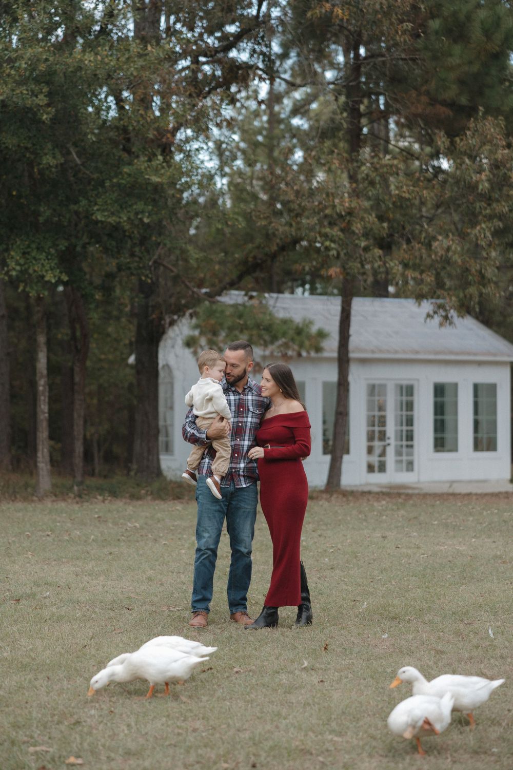 A couple interacts with white geese in a grassy yard outside a white house.