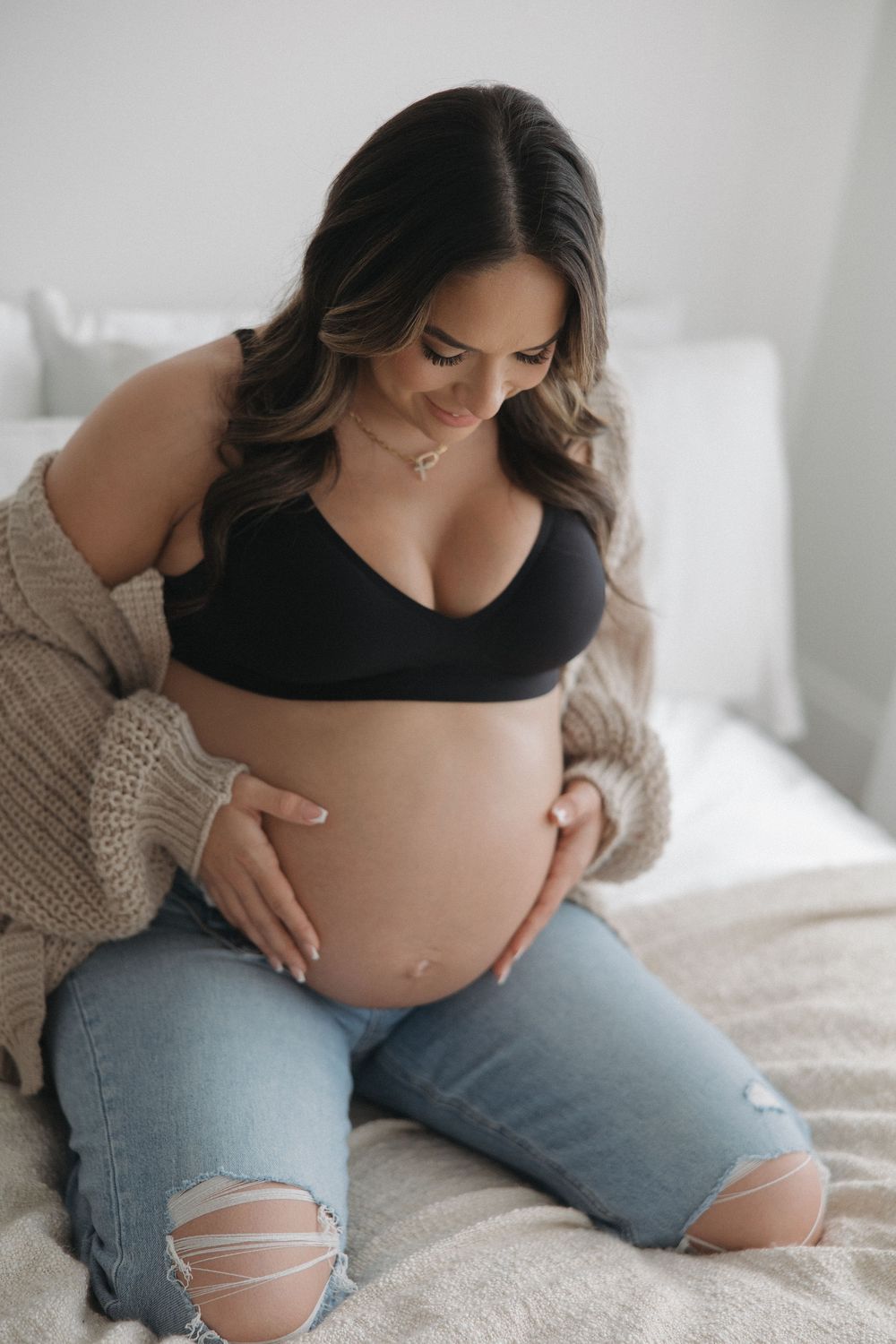 Pregnant woman in ripped jeans and black top sits on white bedding in a series of intimate maternity photos.