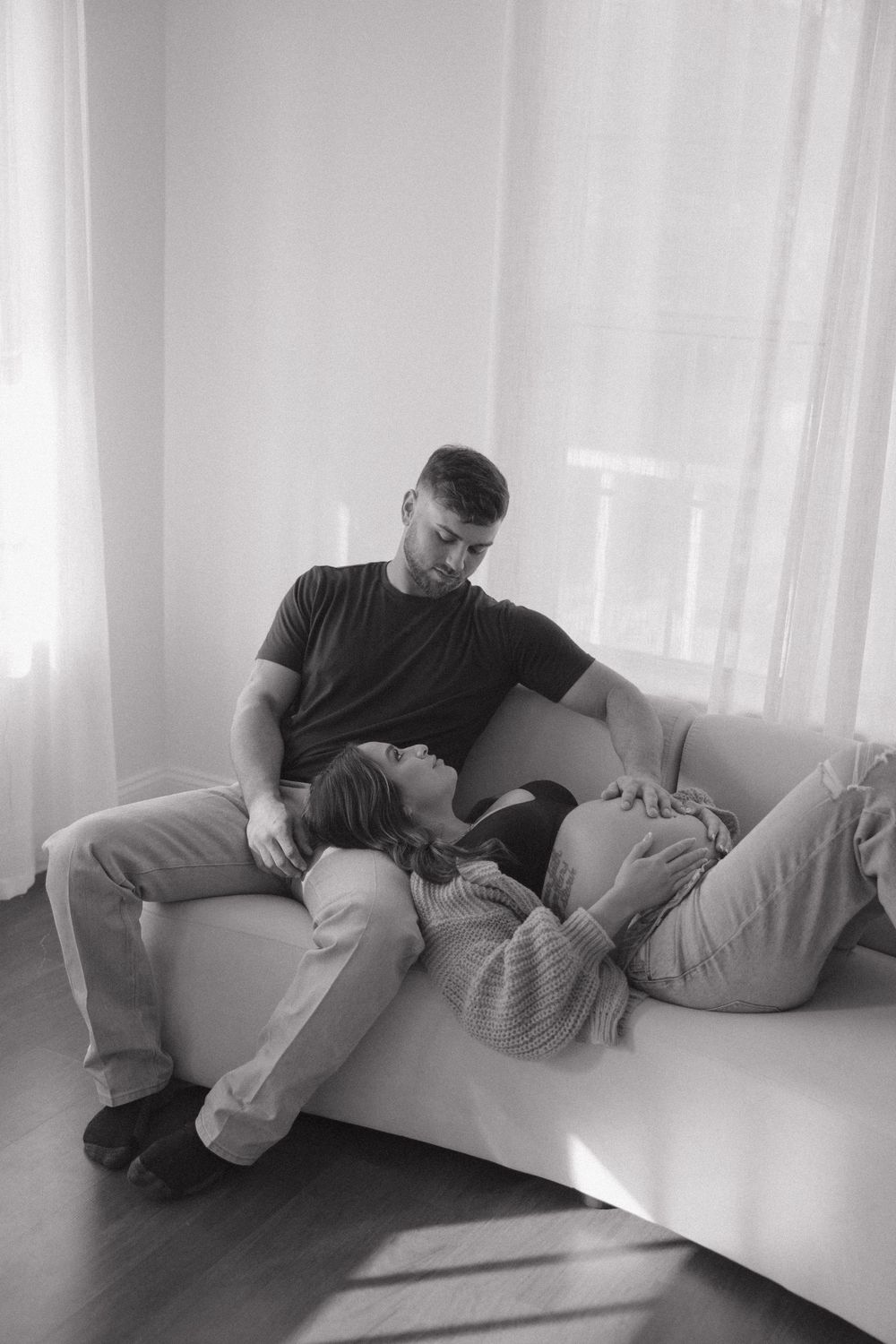 A tender black and white moment of two people resting together on a white couch in a sunlit room.