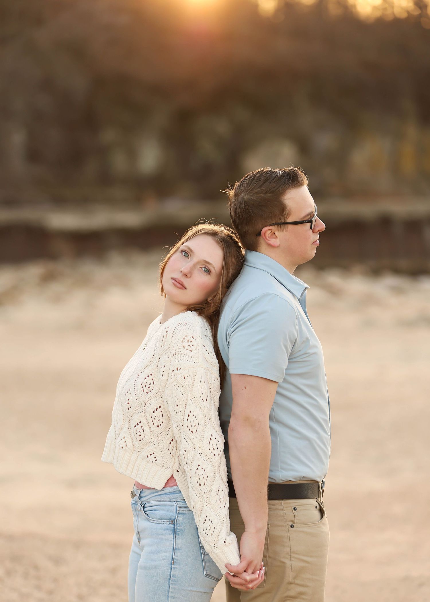 A romantic sunset portrait of a couple in casual attire holding hands back-to-back in a golden field.