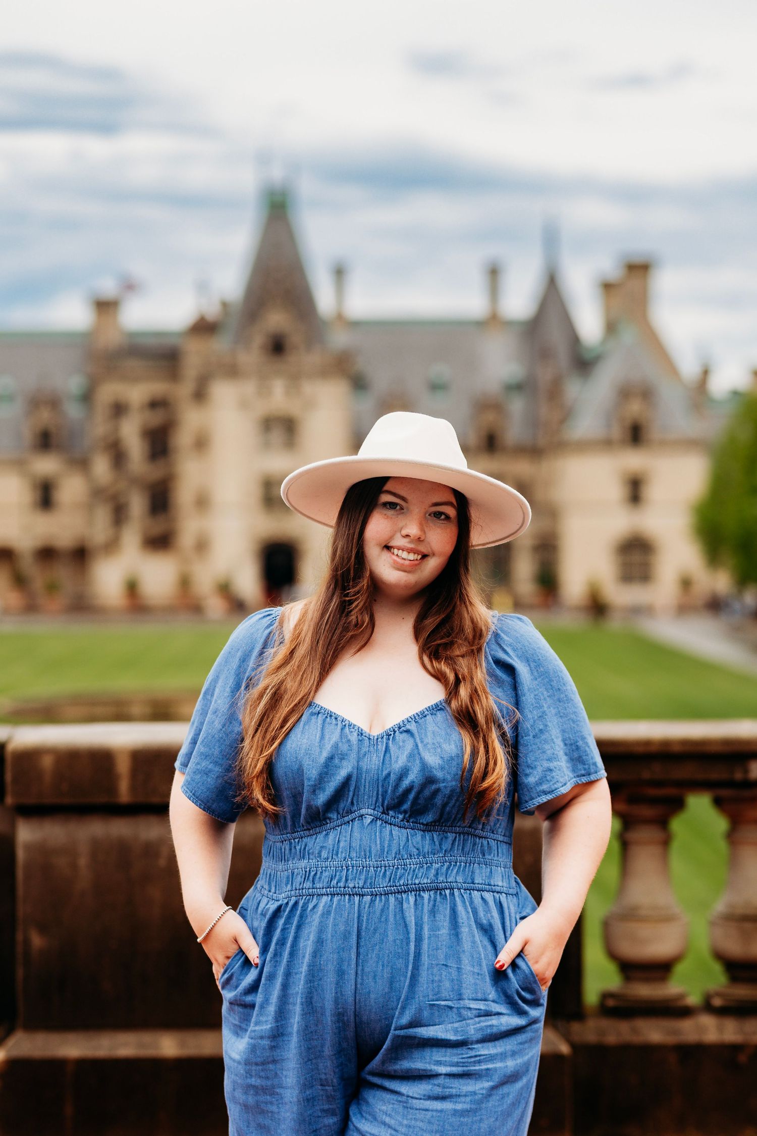 Smiling young woman in blue jumpsuit and white hat poses in front of the Biltmore Estate on a cloudy day.
