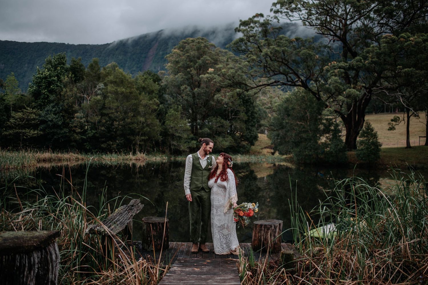 A couple in wedding attire stands on a scenic coastal clifftop overlooking a curved beach and blue ocean.