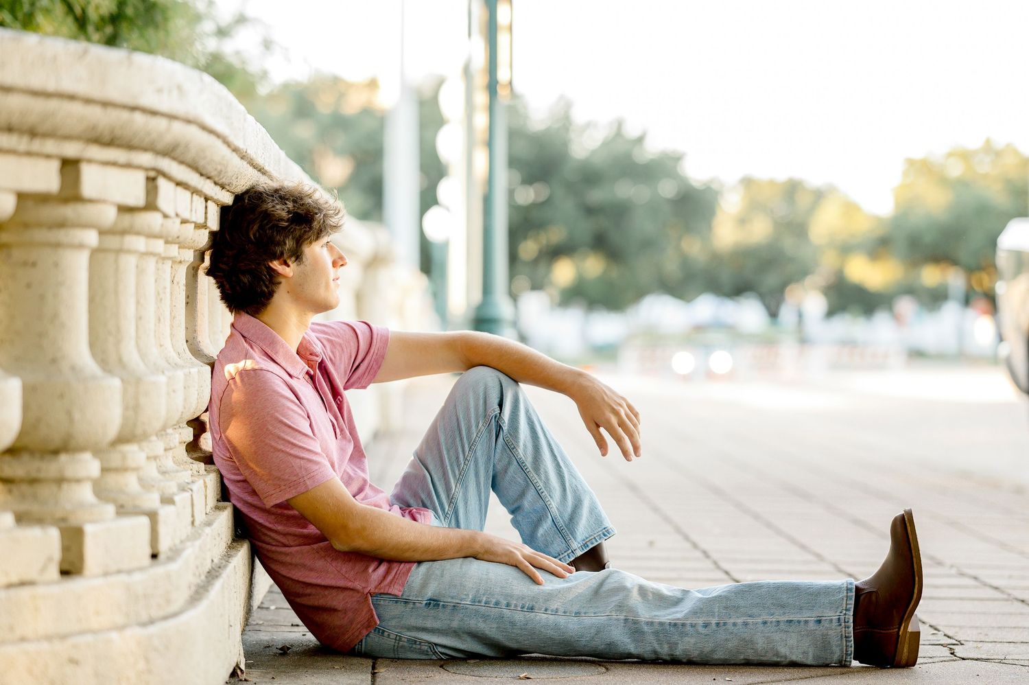 Young man in pink shirt and jeans sitting relaxed against stone columns on an urban sidewalk.