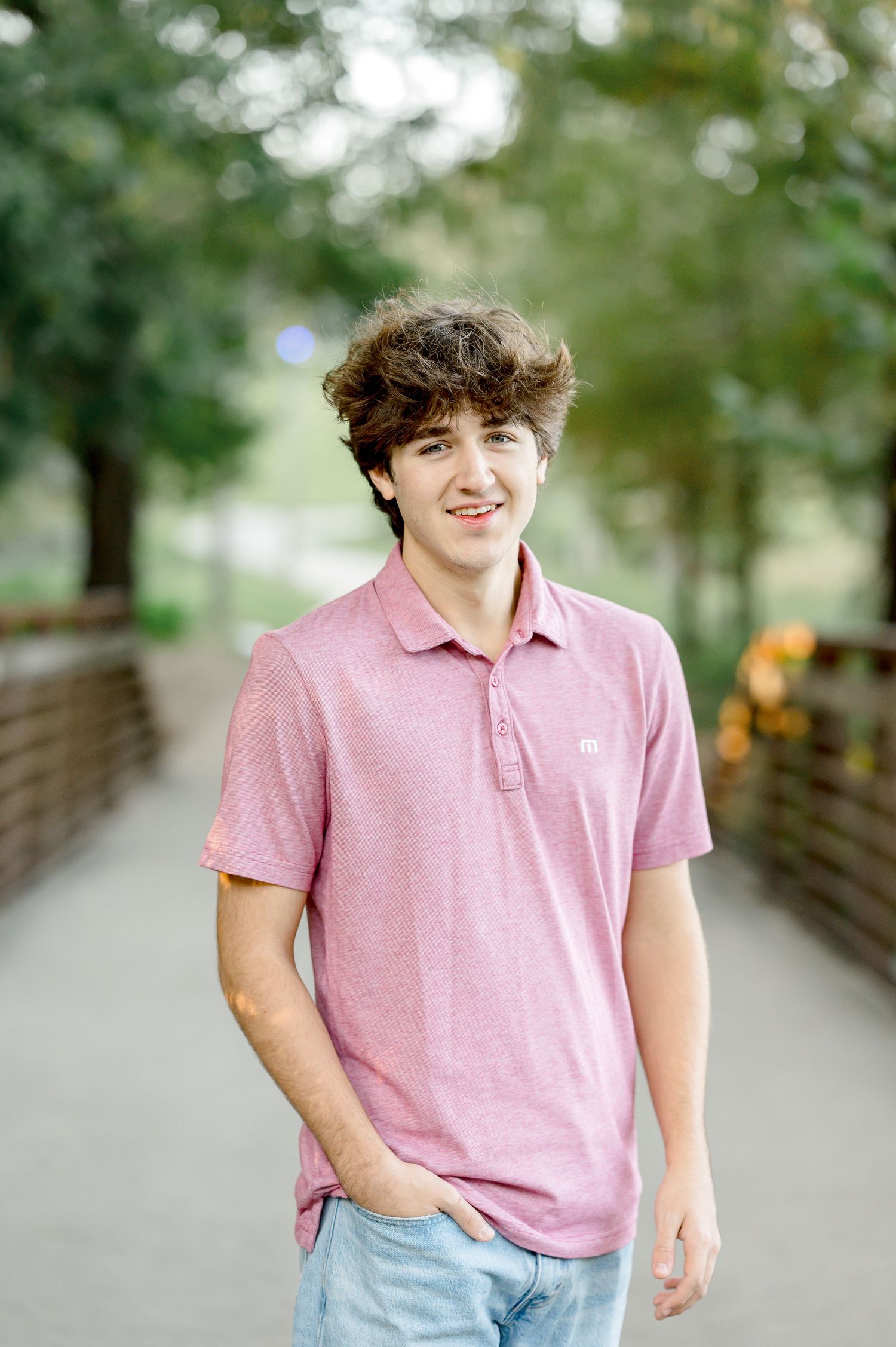 Smiling teenage boy in pink polo shirt standing on a tree-lined pathway, posing for senior portrait photos.