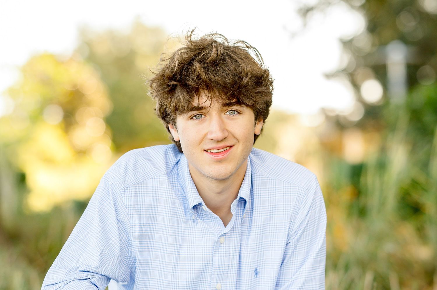 Smiling young man with curly brown hair wearing a light blue shirt outdoors with green foliage background.