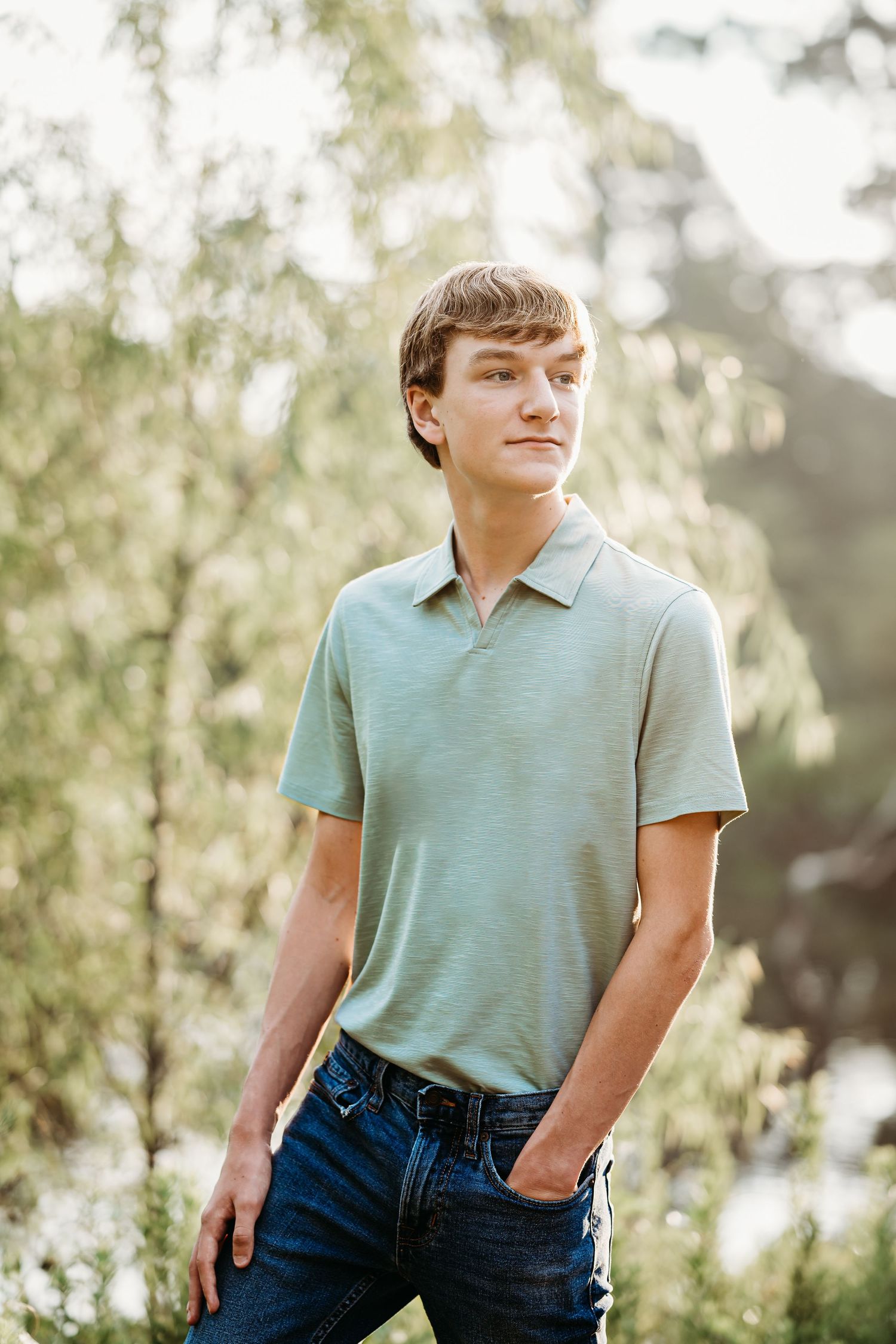 Young man in mint polo shirt and jeans standing outdoors with green trees in background.