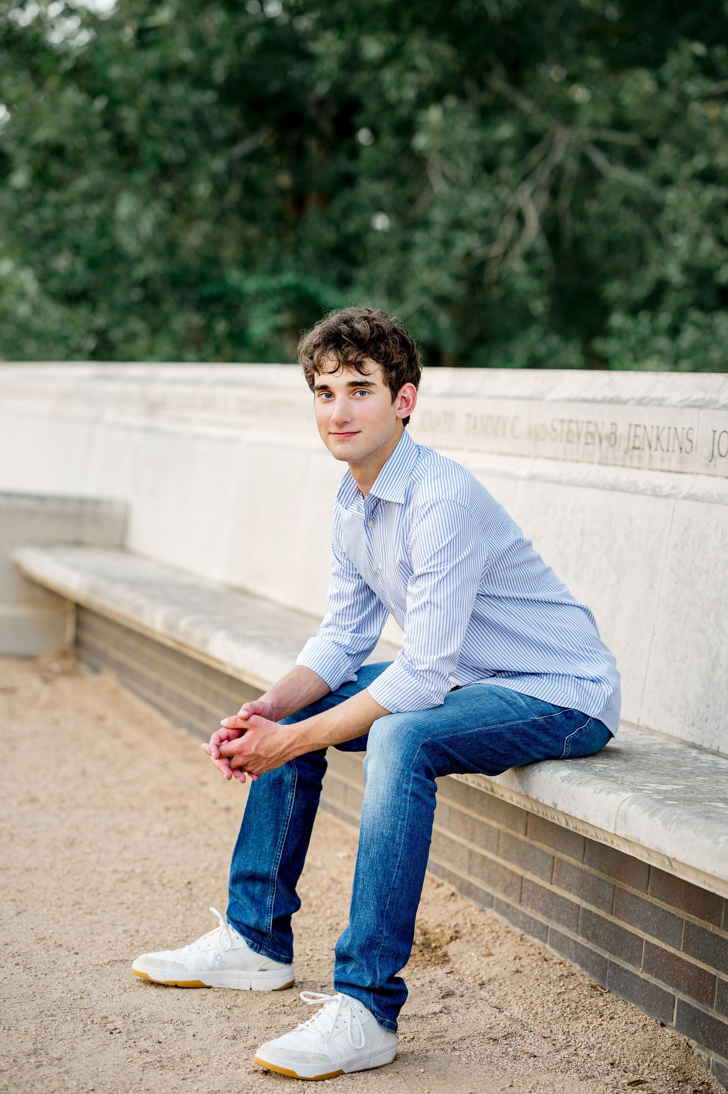 Young man in light blue shirt and jeans sits on stone ledge outdoors, smiling with hands clasped.