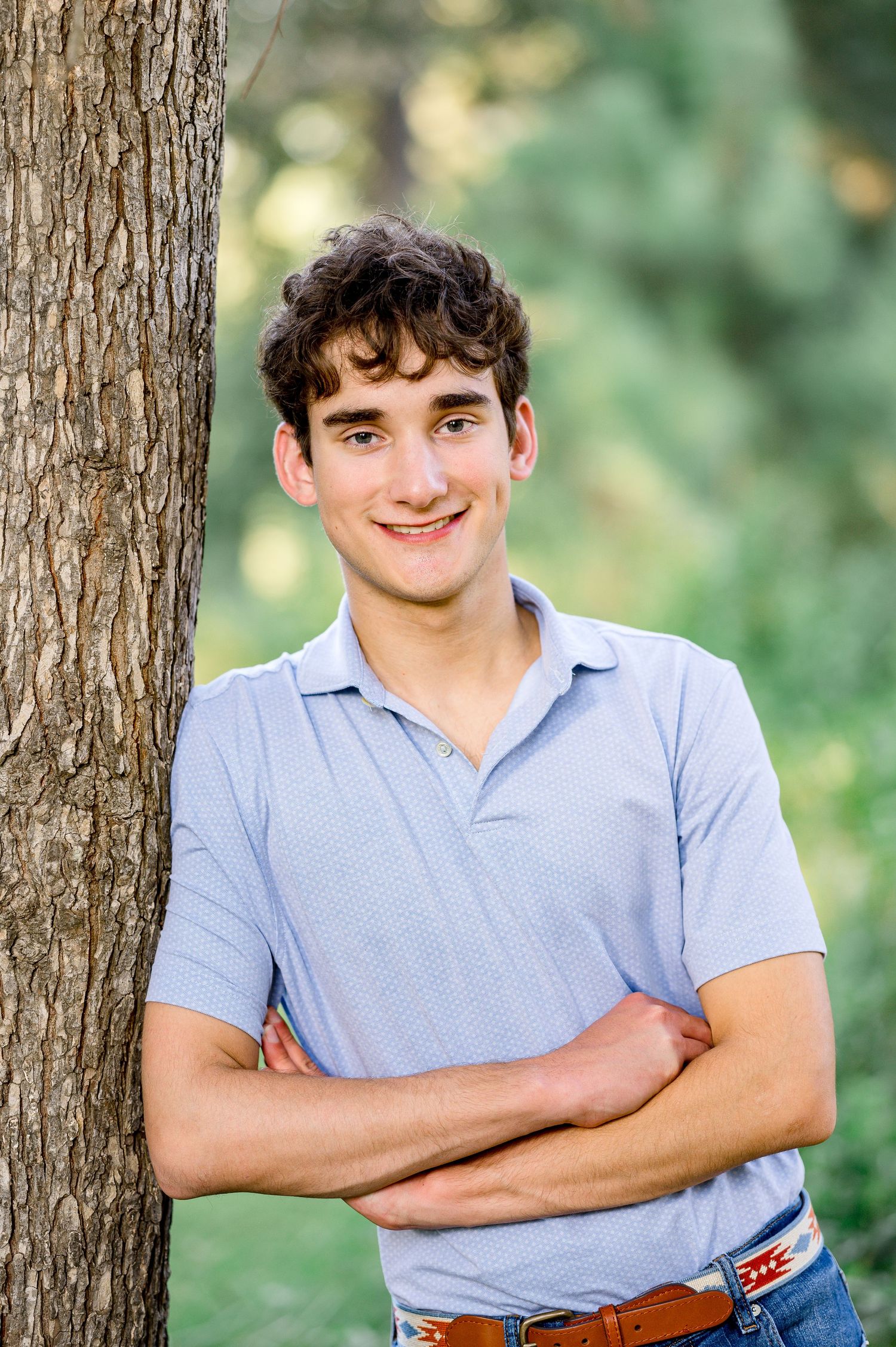 Smiling young man with curly hair leaning against a tree trunk with arms crossed in an outdoor setting.