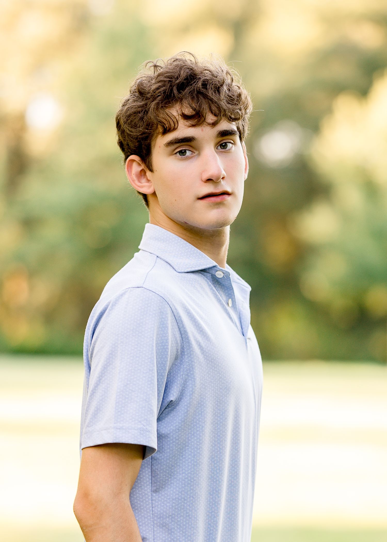 A young man with curly brown hair wearing a light blue polo shirt poses outdoors with a blurred green background.