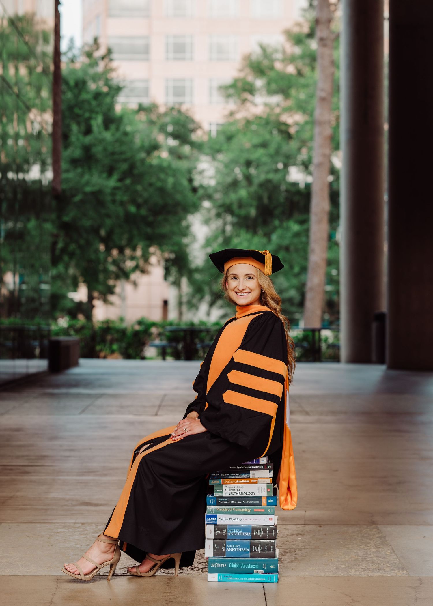 Woman in black and orange doctoral graduation gown and cap sits atop stack of books outdoors.