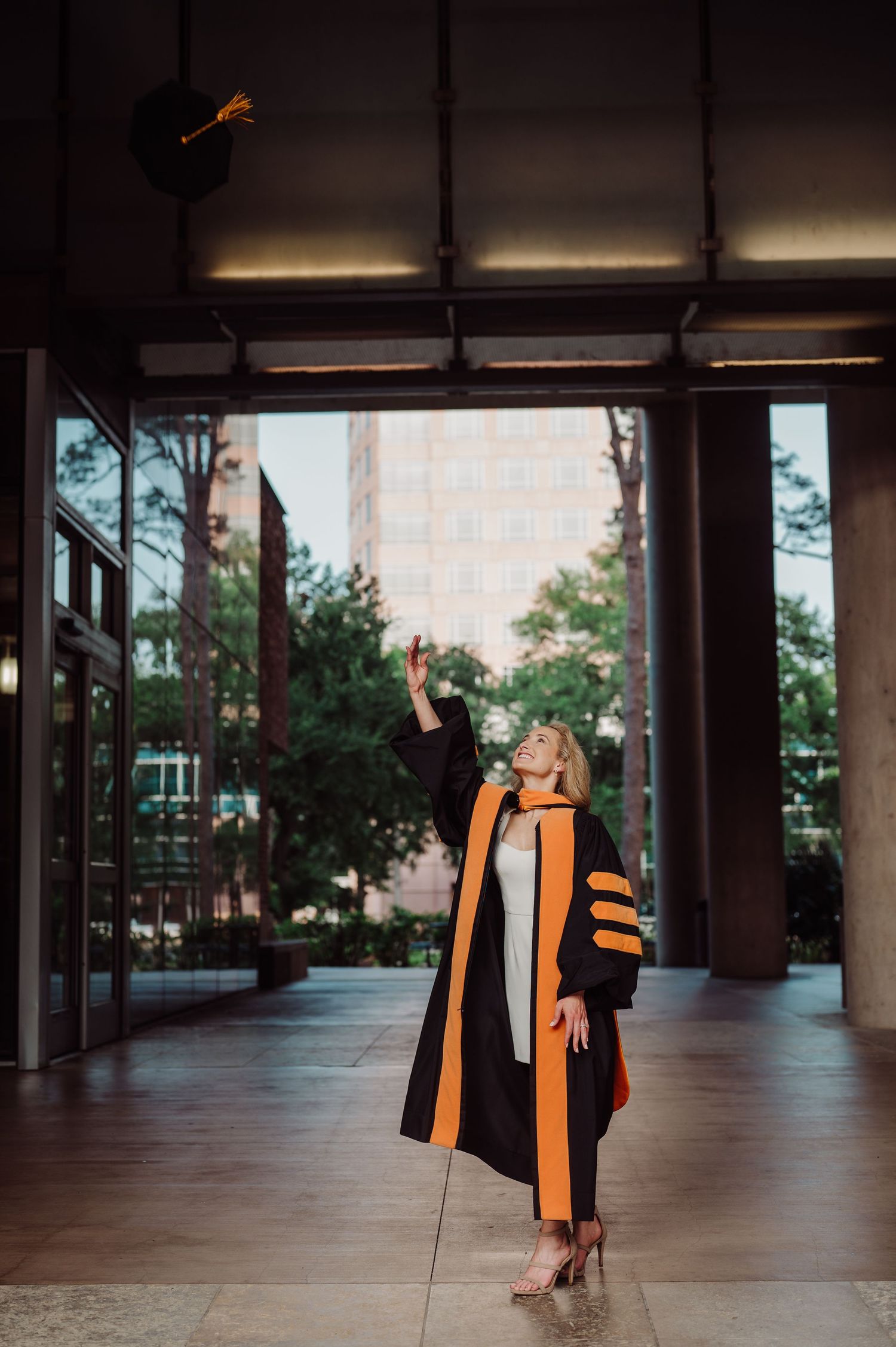 A graduate in black and gold academic regalia tosses her cap in the air inside a modern building corridor.