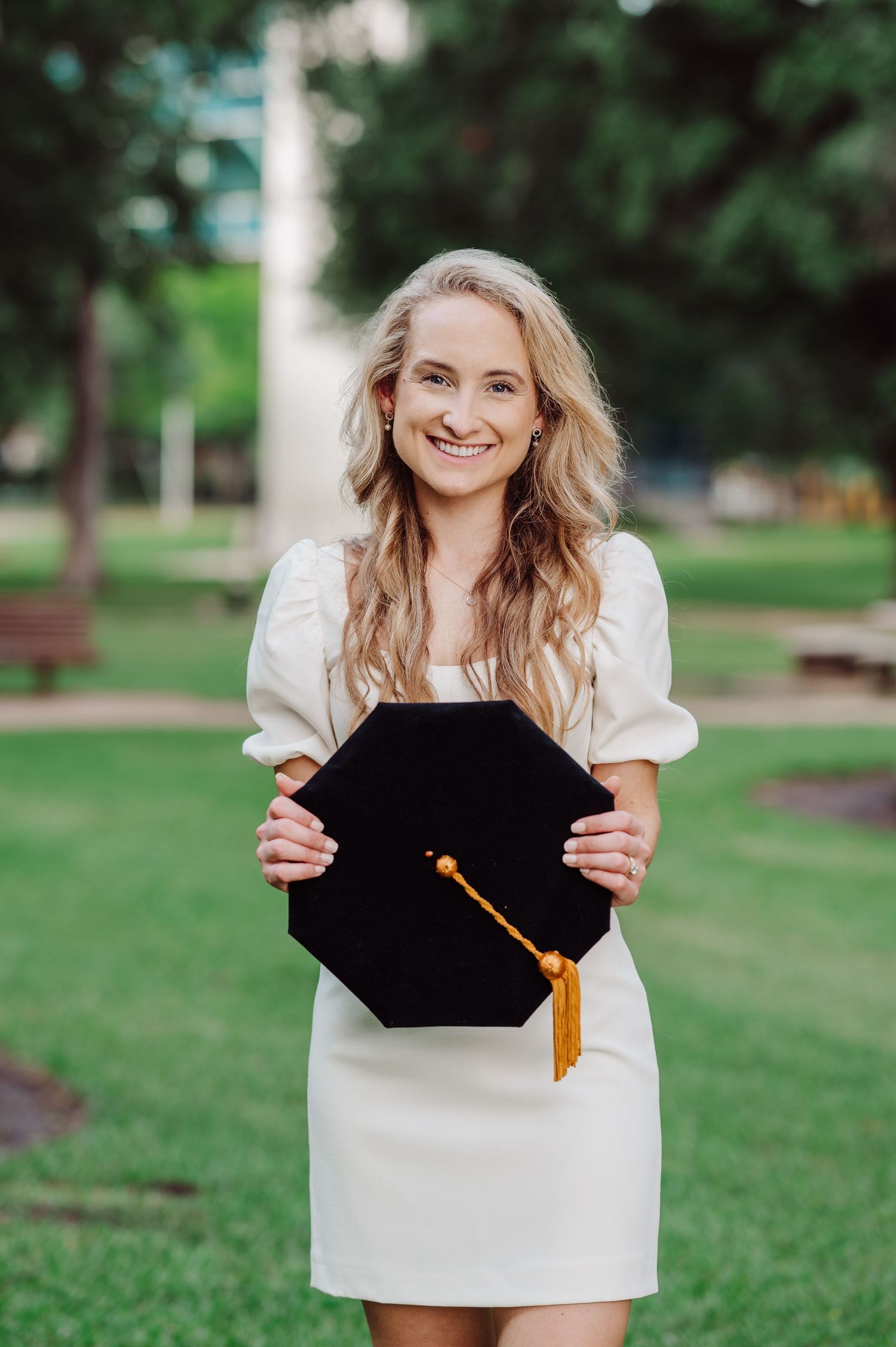 Smiling blonde woman in white dress holding black graduation cap with gold tassel outdoors on green campus.