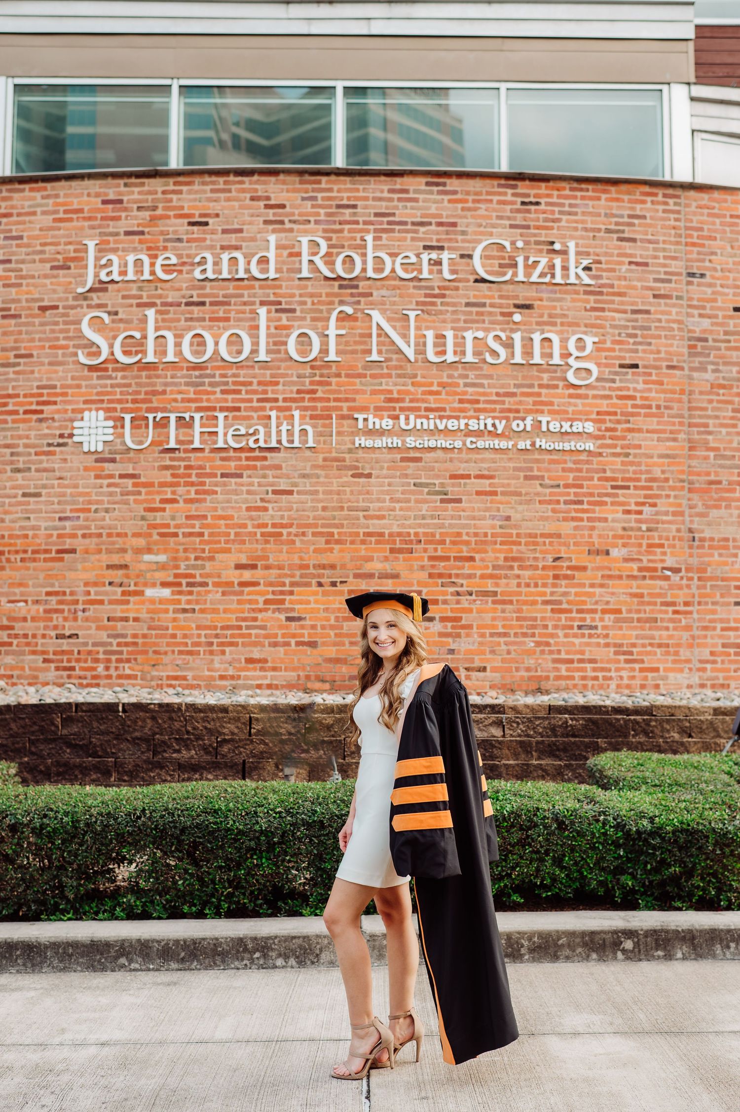 Graduate in cap and gown poses proudly outside Jane and Robert Cizik School of Nursing at UTHealth Houston.