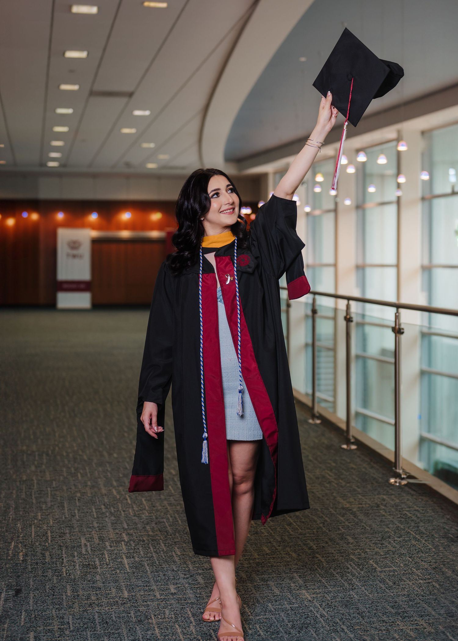 Smiling graduate in black robe raises cap triumphantly in hallway.