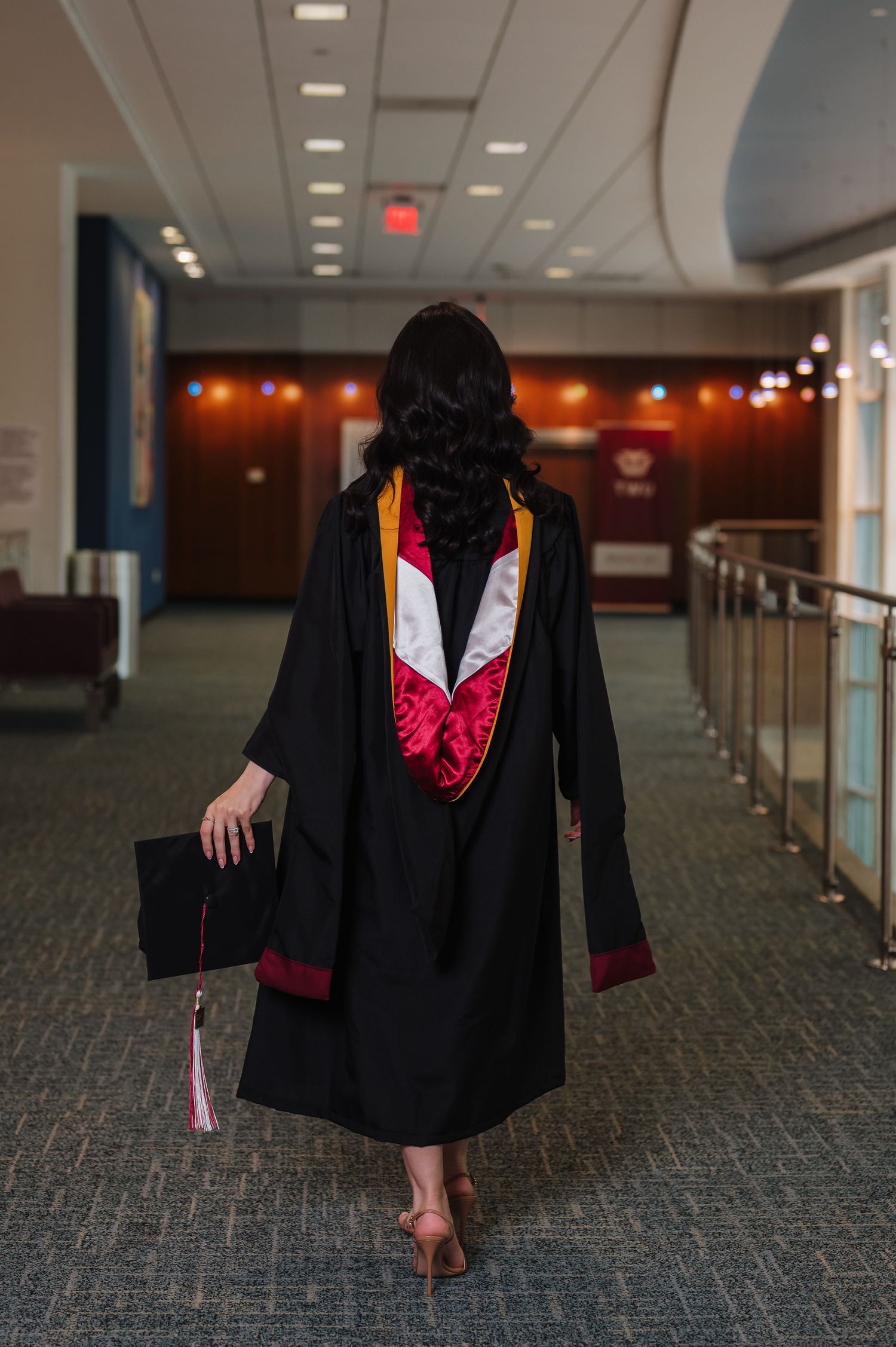 Graduate in black robe walks away down hallway holding cap.