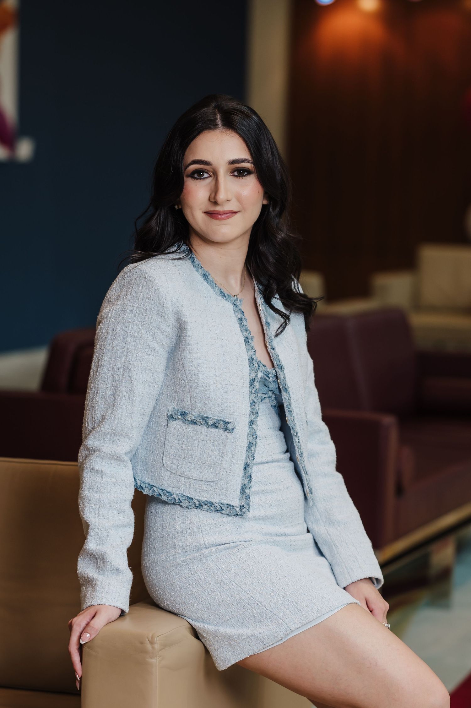 Young woman in light blue tweed jacket and dress sits confidently in a modern interior setting.