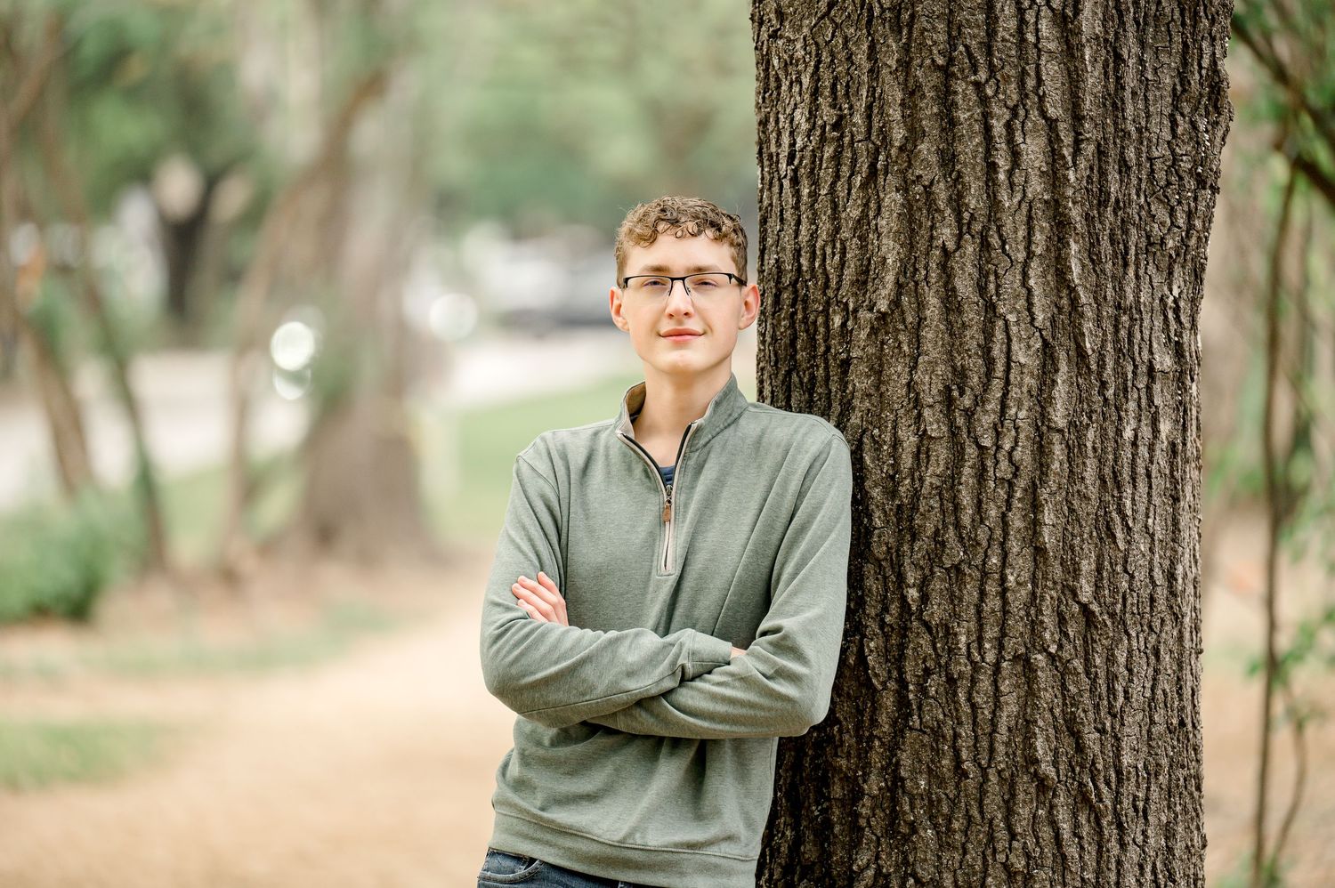 Young man with glasses in gray zip-up jacket leans against tree with arms crossed in a park setting.