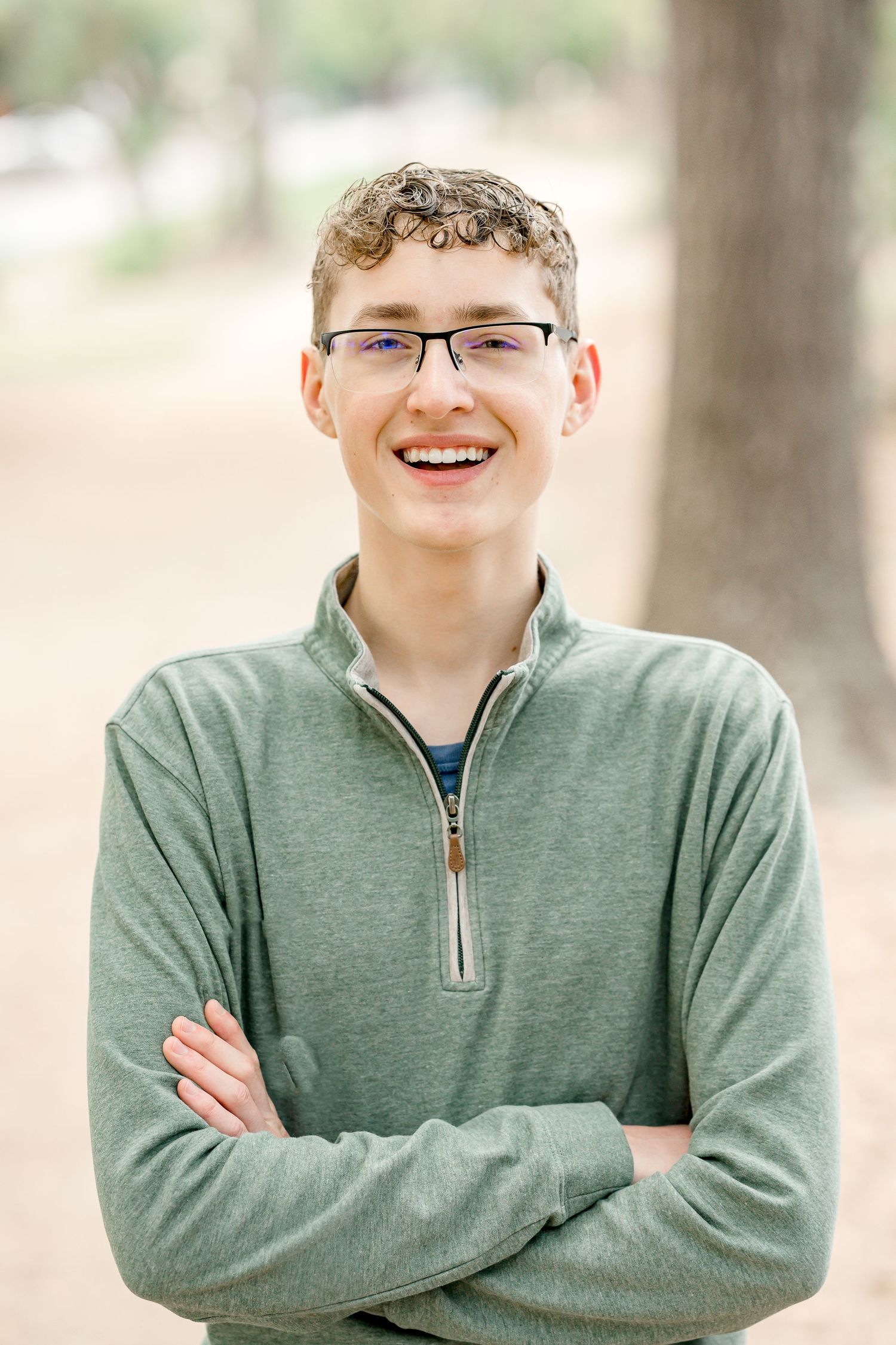 Smiling teen boy with curly hair and glasses wearing a green zip-up sweatshirt with arms crossed outdoors.