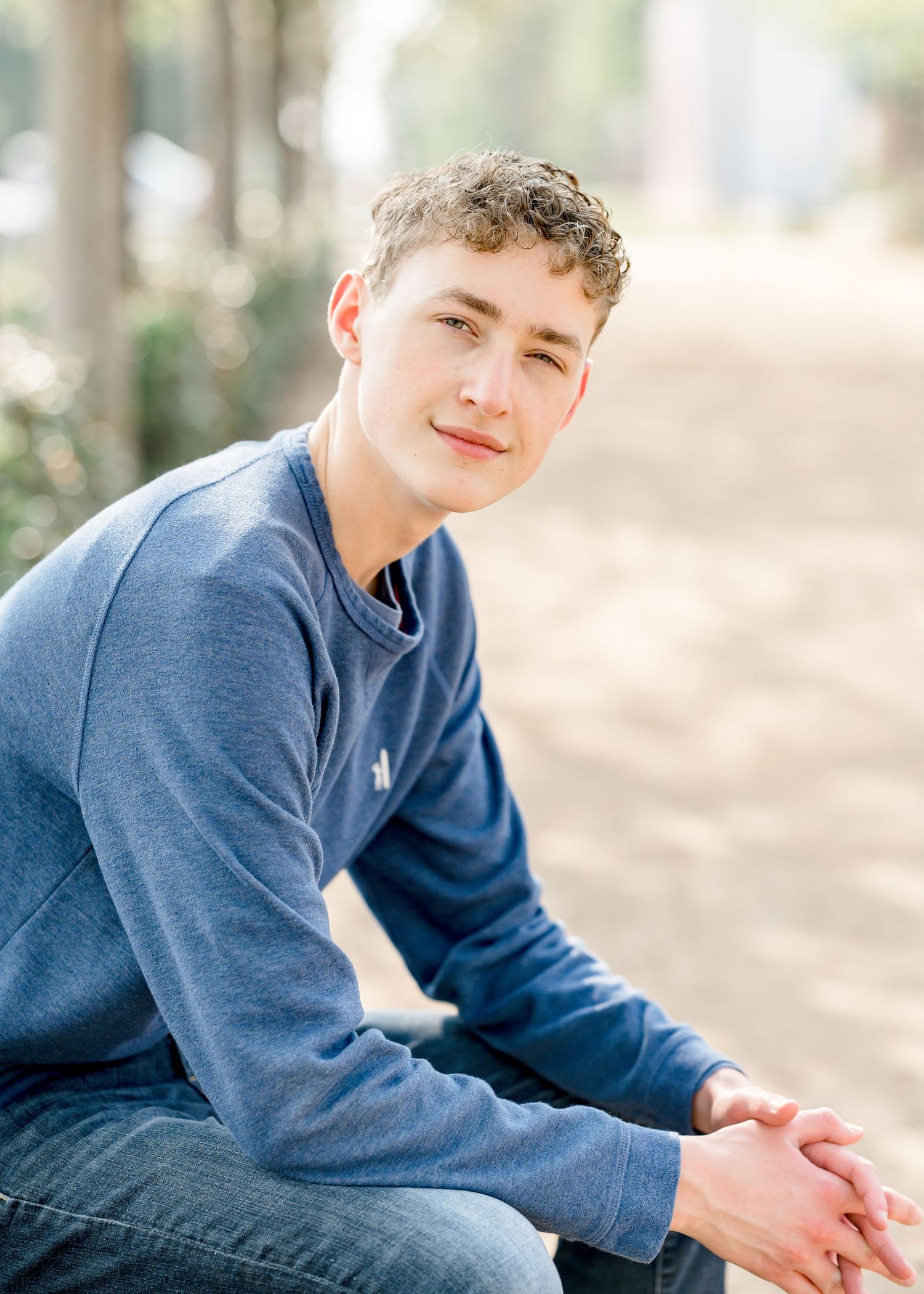 Teen boy with curly hair in a blue sweatshirt sitting outdoors, looking over his shoulder with a relaxed expression.