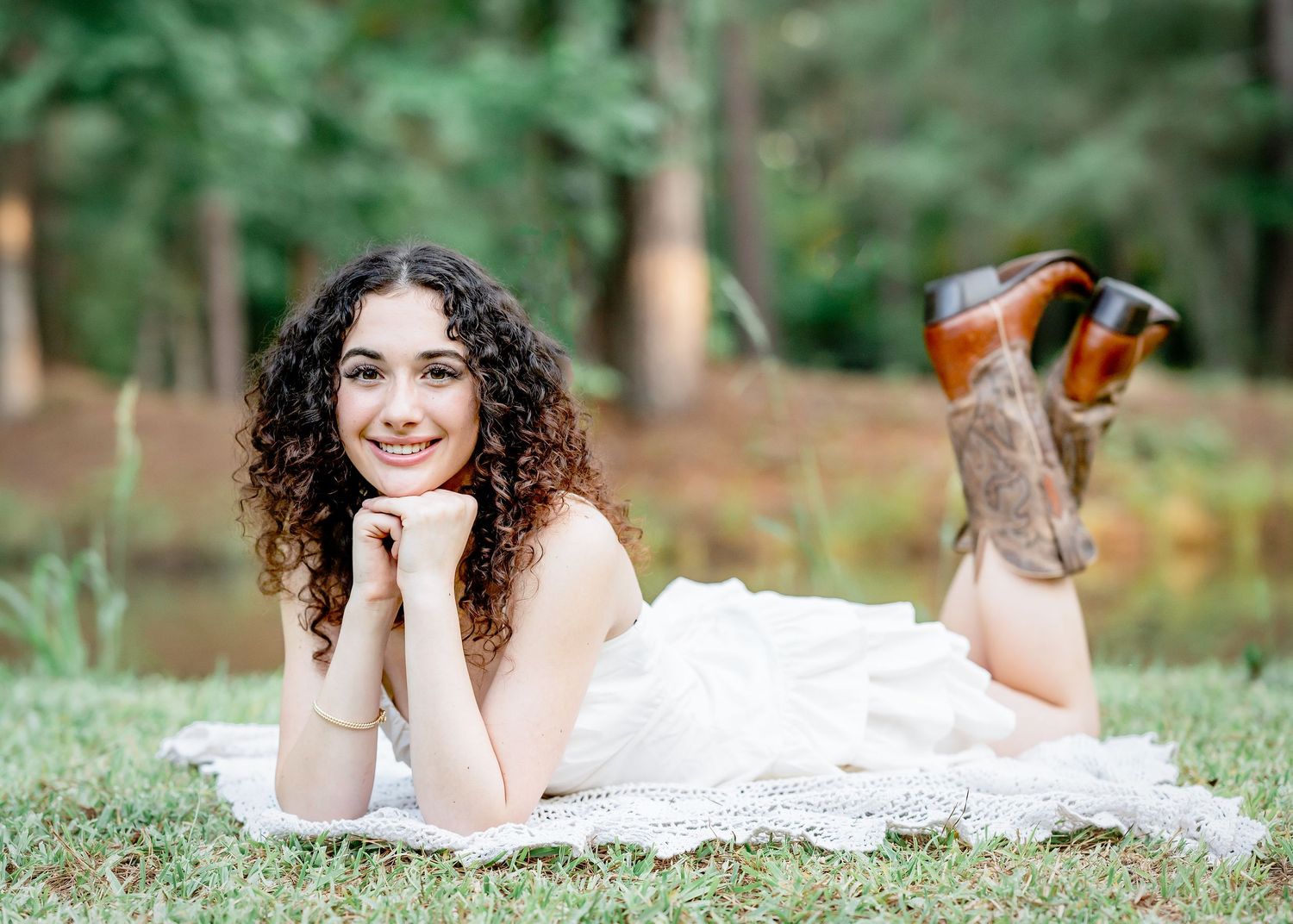 Young woman with curly hair in white dress and cowboy boots lying on grass, smiling outdoors.