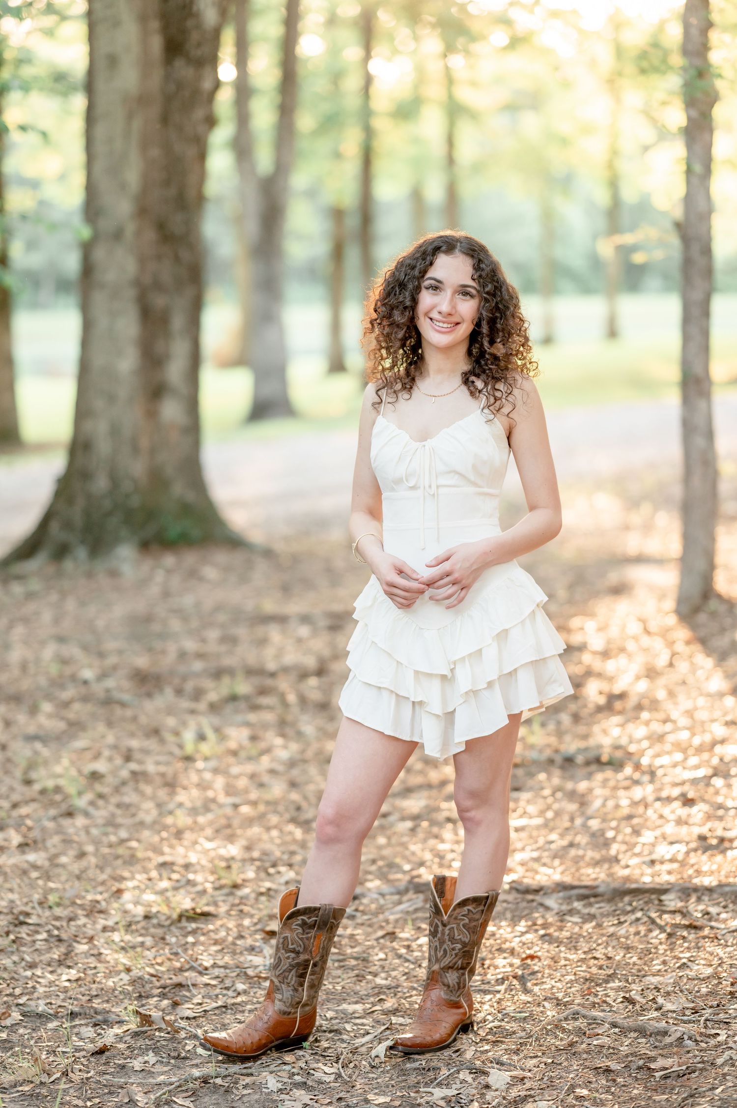 Young woman with curly hair in white ruffled mini dress and brown cowboy boots standing among trees outdoors.