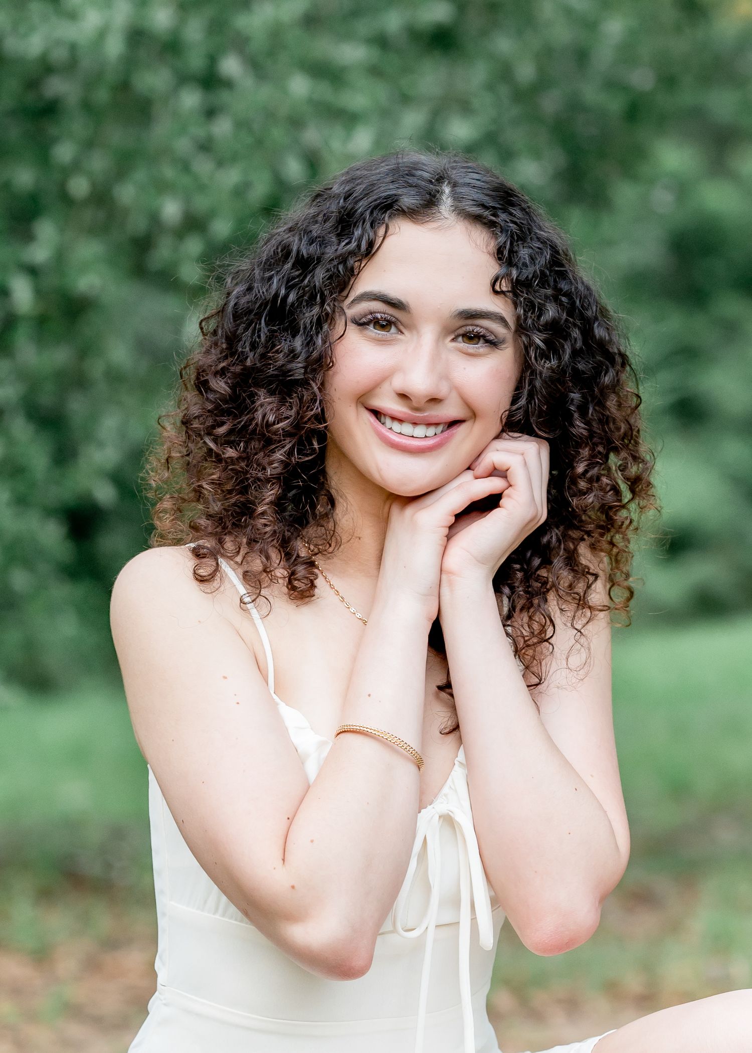 Young woman with curly hair smiling warmly, hands clasped near face, wearing white dress outdoors.