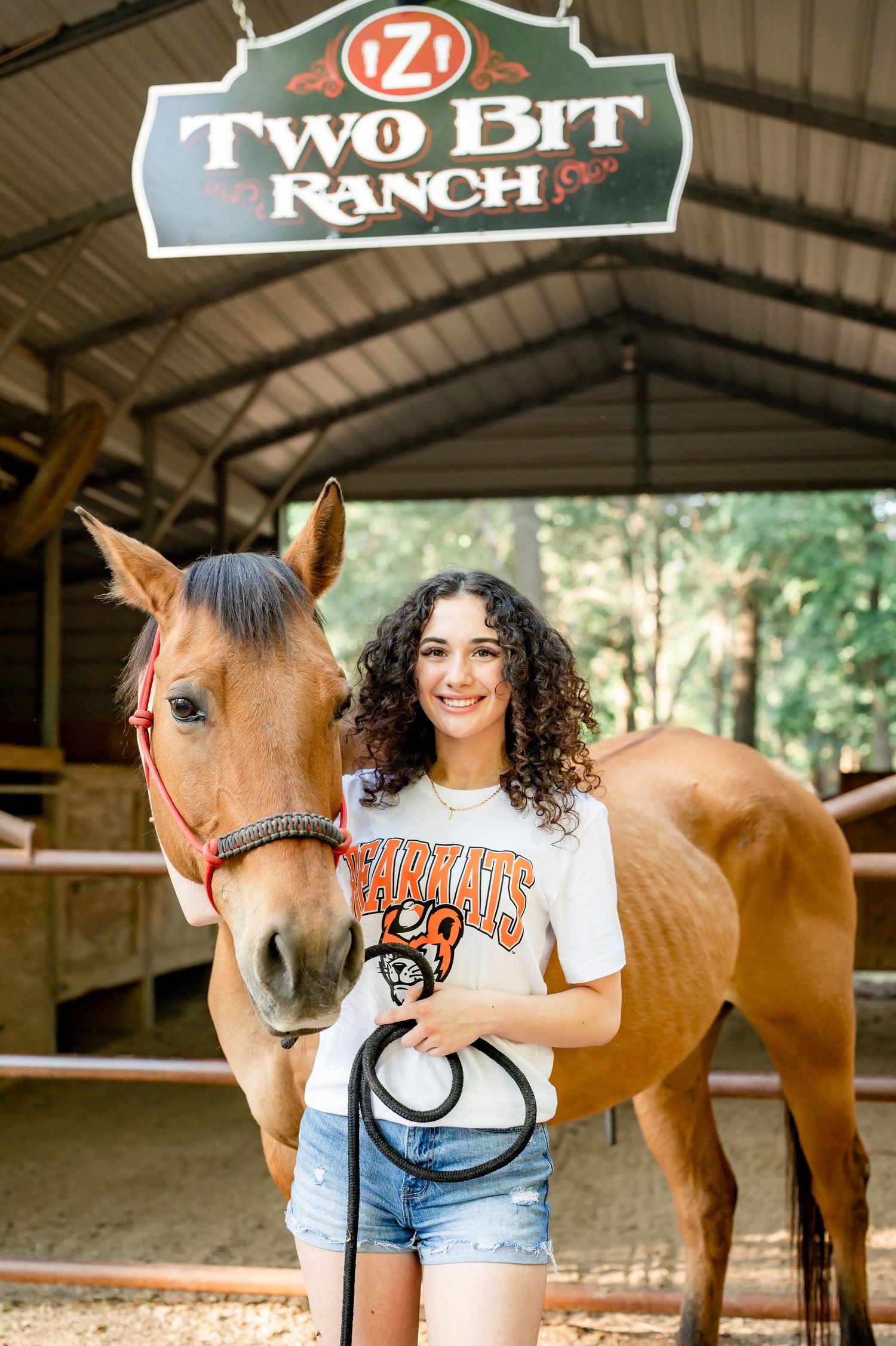 Young woman with curly hair holding a horse's lead rope at Two Bit Ranch stables, wearing a Bearkats shirt and denim shorts.