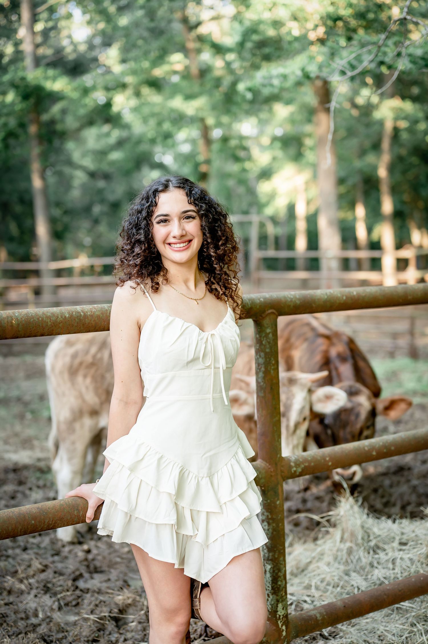 Young woman in white ruffled dress smiling by a rustic fence with brown cattle and green trees in background.