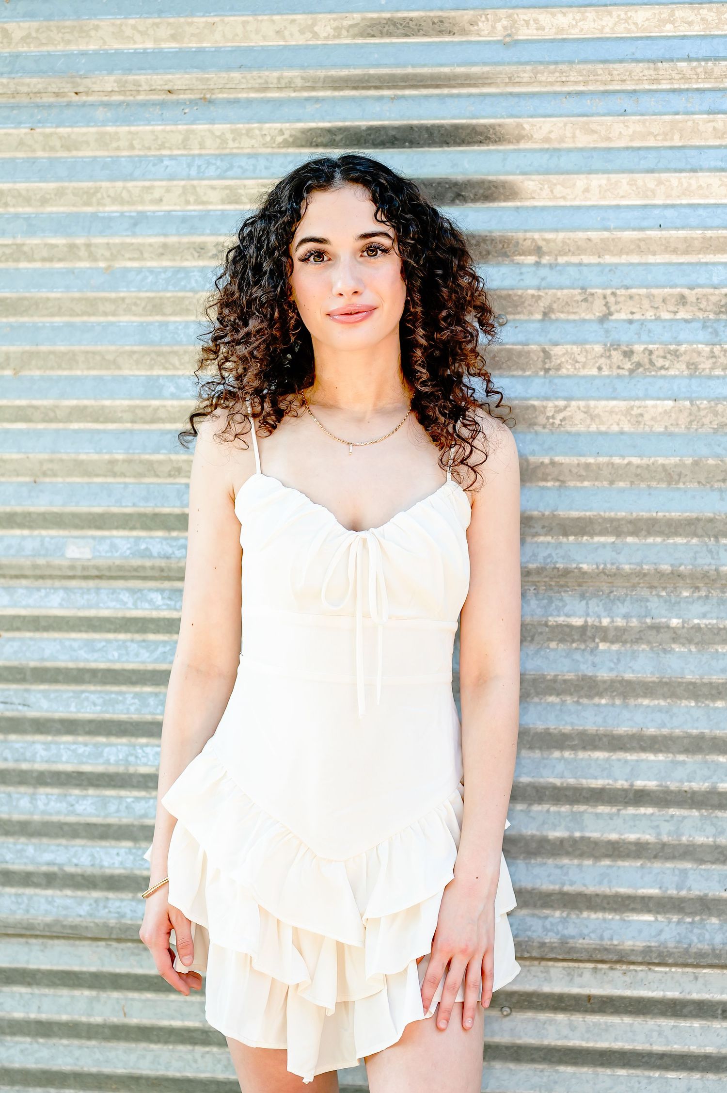 Young woman with curly hair wearing a white ruffled mini dress, posing against a corrugated metal wall.