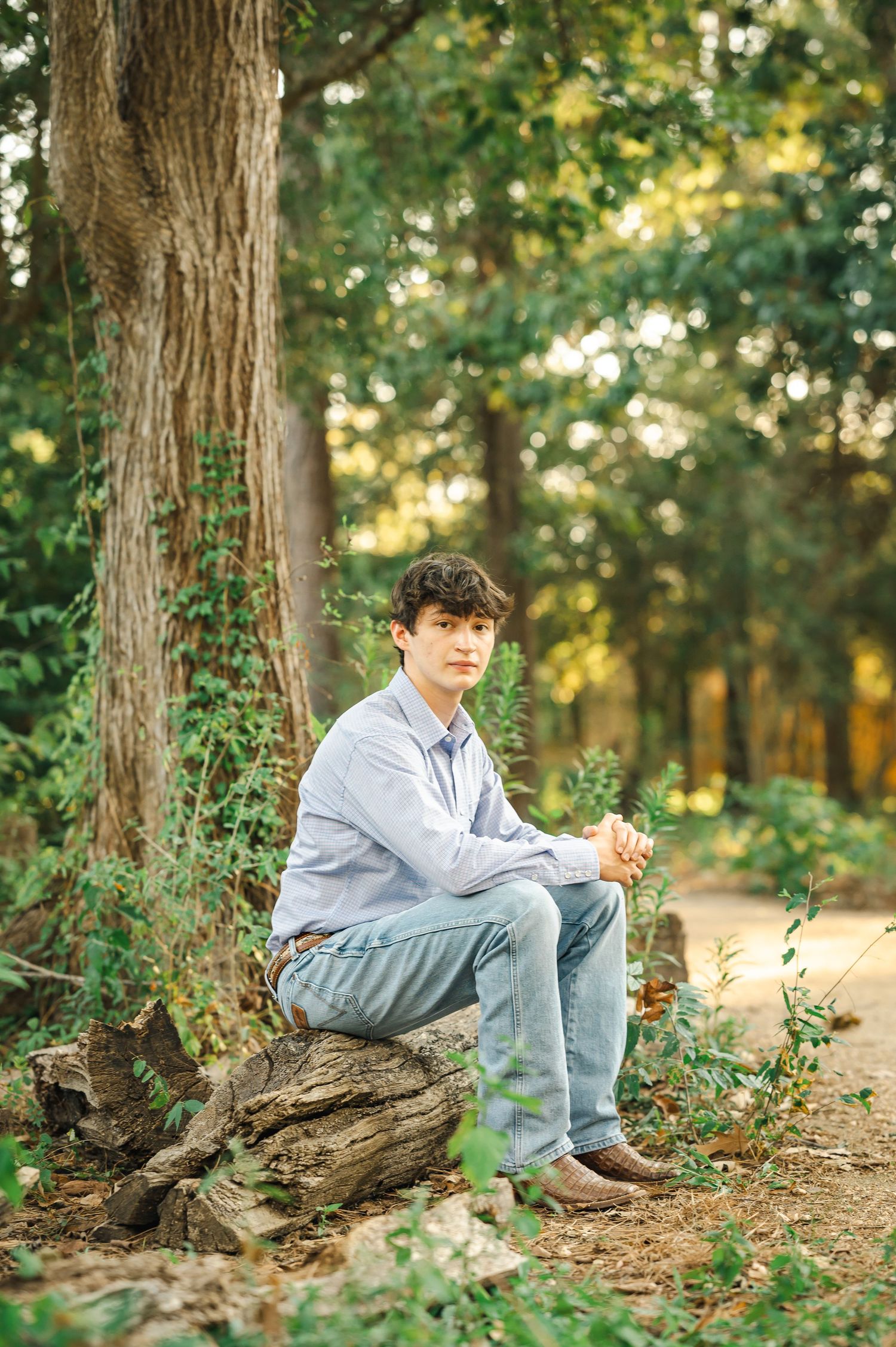 Young man in blue shirt and jeans sitting on a log in a lush green forest setting.