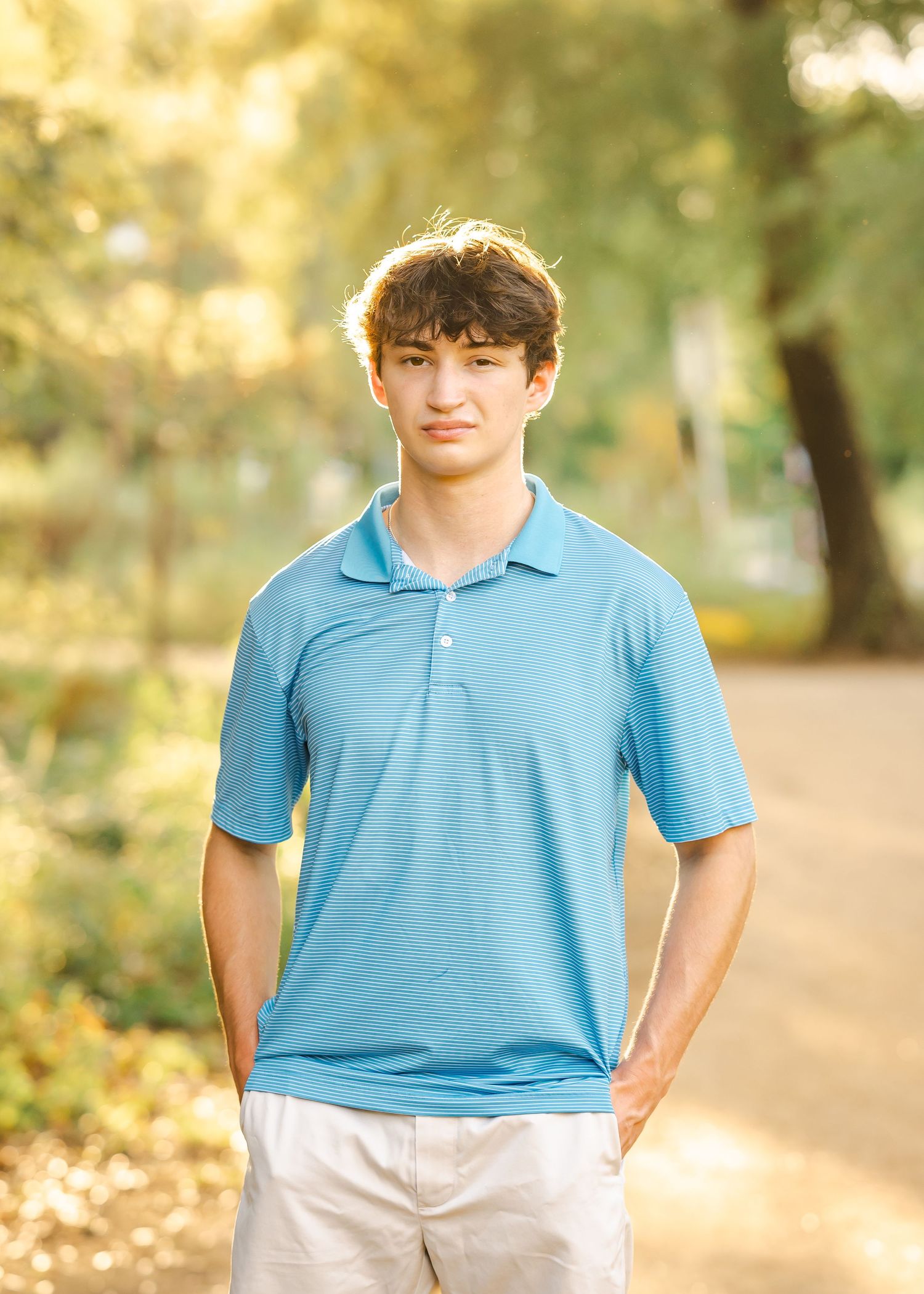 A young man in a blue polo shirt and khaki shorts stands confidently outdoors with a blurred green park background.