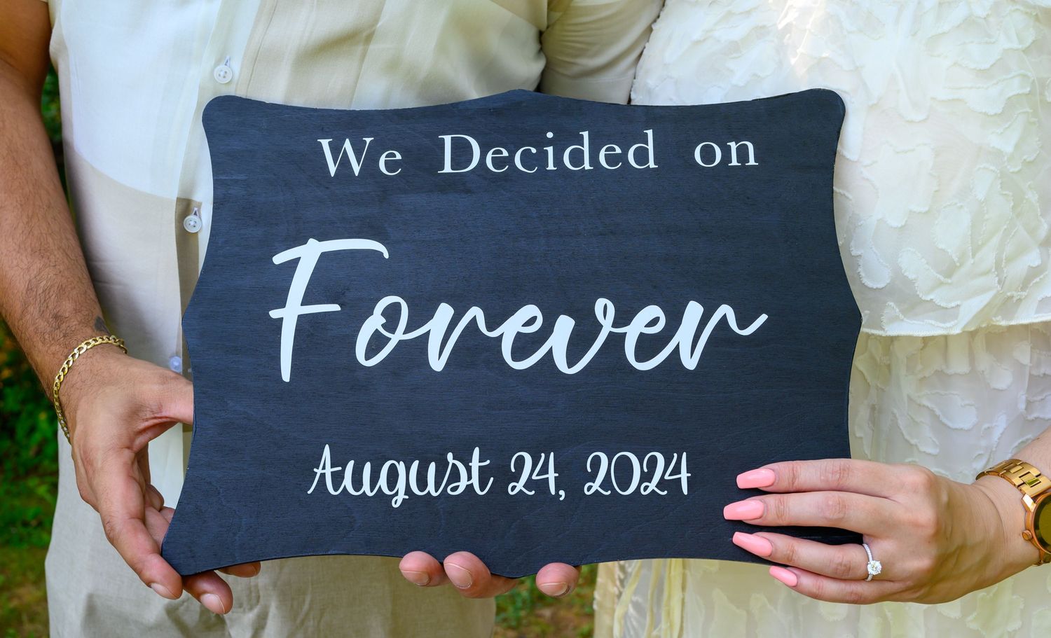 Person holds a dark slate sign with 'We Decided on Forever' and wedding date August 24, 2024 written in white text.