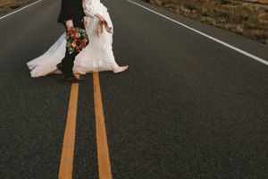 Bride and groom crossing road in wedding attire with colorful bouquet on open highway.