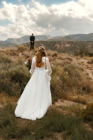 Bride in backless white gown approaching groom standing at desert overlook with bouquet.