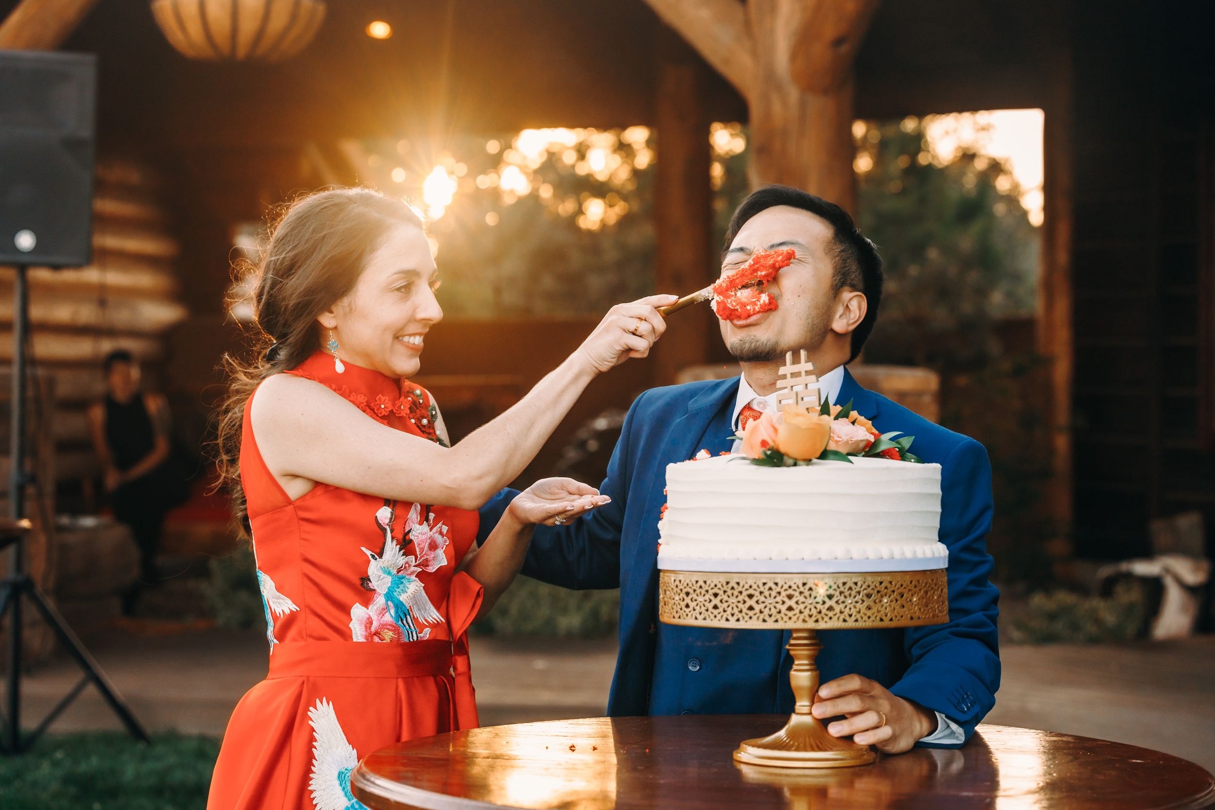 Bride in red qipao feeds groom cake slice at wedding reception with white tiered cake.