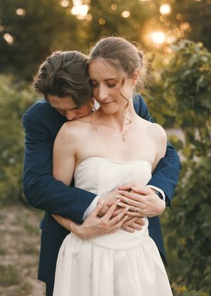 A groom in a navy suit embraces his bride in a white strapless gown during a golden hour outdoor wedding portrait.