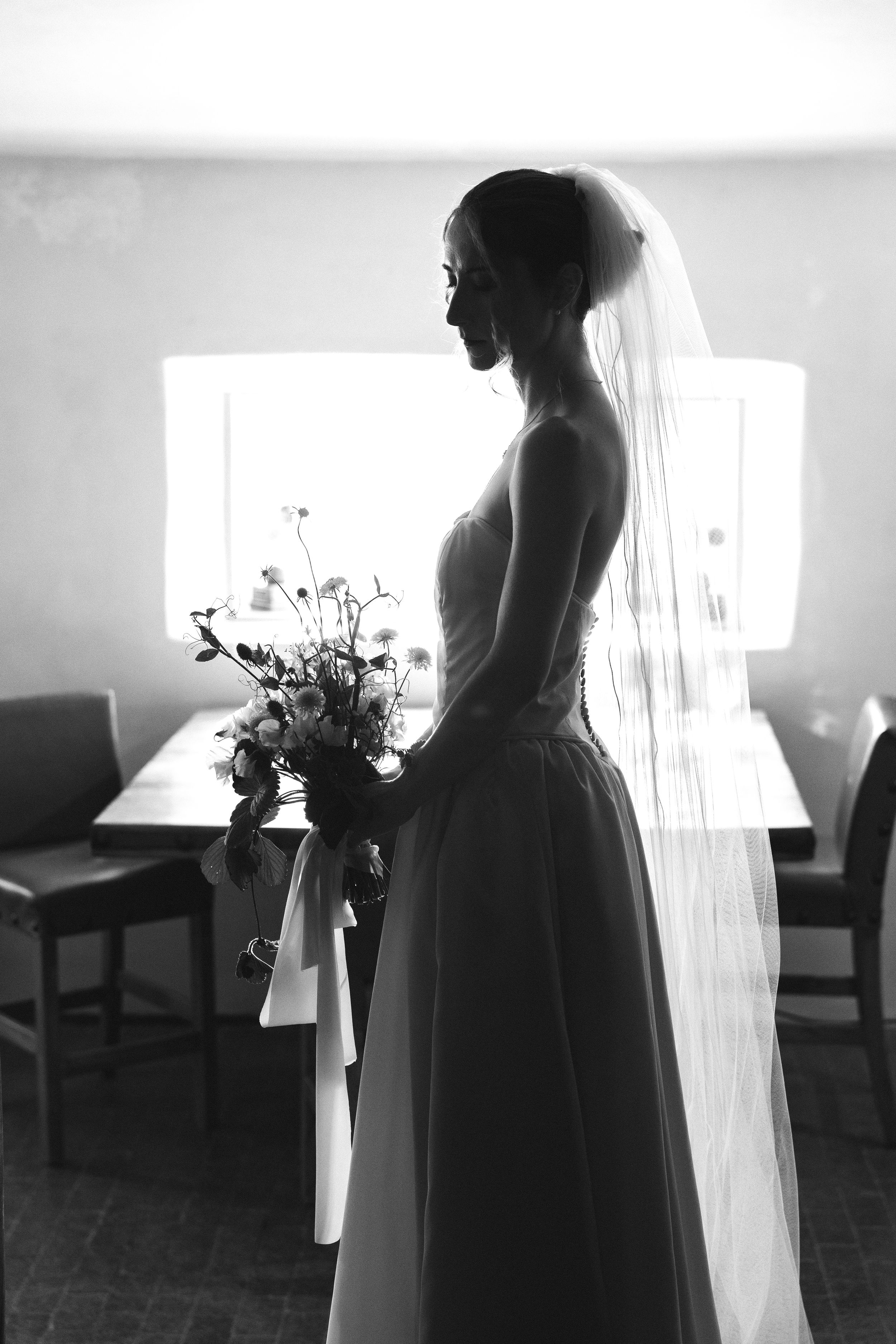 Black and white silhouette of a bride in a veil holding a bouquet, standing by a window in soft light.