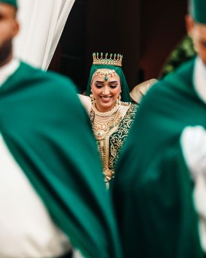 A smiling bride in green and gold traditional attire with a crown headpiece, framed by green fabric drapes.