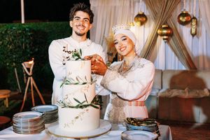 Muslim couple in wedding attire cutting a white floral cake at an outdoor reception celebration.