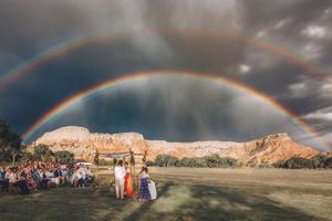 Ghost Ranch Wedding ceremony under a stunning double rainbow over red rock canyon cliffs with guests seated on green lawn.