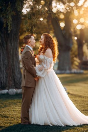 Bride in off-shoulder white tulle gown and groom in tan suit embrace at golden sunset under large trees with string lights.