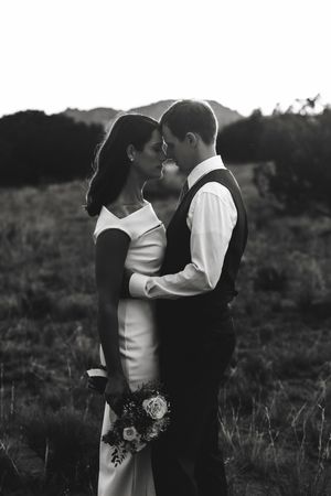Black and white portrait of bride and groom nearly kissing in field with mountain backdrop.