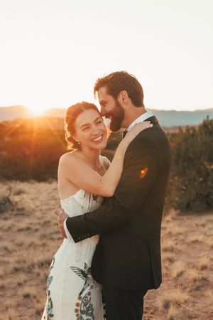 A smiling bride embraces her bearded groom at golden hour in a desert landscape.