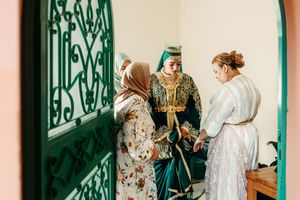 Three women in traditional Moroccan attire gather near an ornate green door during a wedding ceremony.